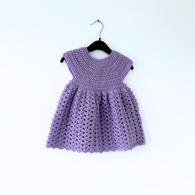 May include: A purple crocheted baby dress with a scalloped hem and short sleeves. The dress is hanging on a black hanger against a white background.