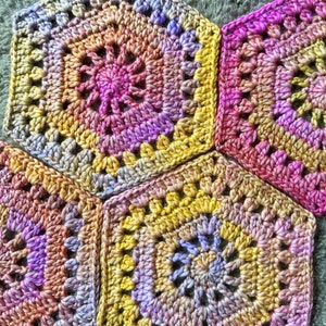 May include: Three crocheted granny squares in shades of pink, yellow, and purple. The squares are joined together to form a larger piece of fabric.