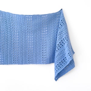 May include: A light blue crocheted shawl with a delicate openwork pattern. The shawl is hanging on a white wall.