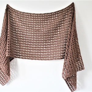 May include: A brown crocheted shawl with a square pattern. The shawl is hanging on a white wall.