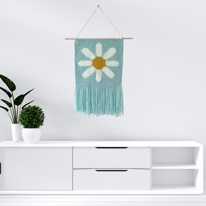May include: A light blue crocheted wall hanging with a white daisy design and a yellow center. The hanging has a fringe border and is attached to a wooden dowel.