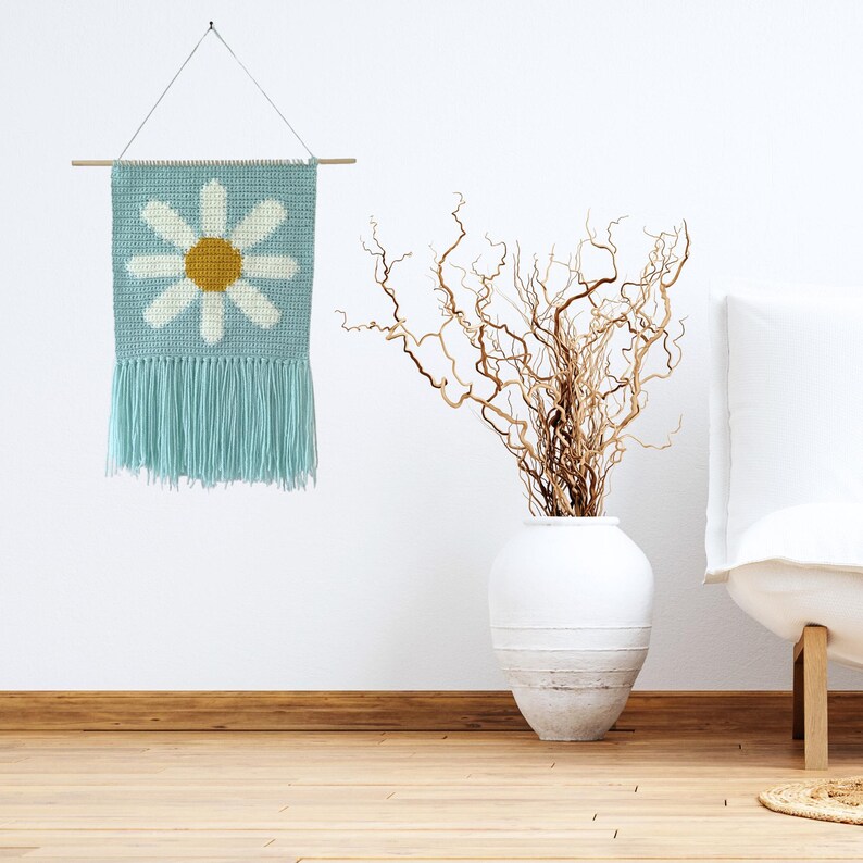 May include: A light blue crocheted wall hanging with a white daisy with a yellow center. The hanging has fringe on the bottom and is hung on a wooden dowel.