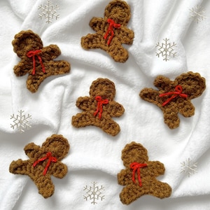 May include: Crocheted gingerbread man ornaments in shades of brown, each with a red ribbon bow. The ornaments are arranged on a white, textured surface with silver snowflake accents, creating a festive holiday theme.