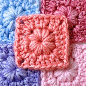 Op de afbeelding: Close-up van kleurrijke gehaakte vierkanten. Het centrale vierkant is koraal met een bloemmotief. Omringende vierkanten zijn blauw, paars, roze en bordeaux, elk met een bloemmotief. Het garen heeft een zacht, getextureerd uiterlijk.