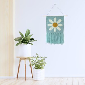 May include: A light blue crocheted wall hanging with a white daisy design and a yellow center. The hanging has a fringe border and is displayed on a white wall with two potted plants.
