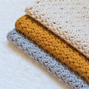 May include: Three crocheted dishcloths in different colours: grey, mustard yellow, and cream. The dishcloths are stacked on top of each other.