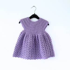May include: A purple crocheted baby dress with a scalloped hem and short sleeves, hanging on a black hanger against a white background.