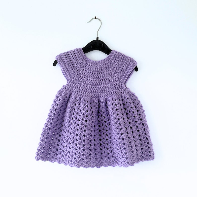 May include: A purple crocheted baby dress with a scalloped hem and short sleeves, hanging on a black hanger against a white background.