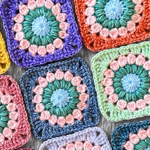 May include: Close-up of colourful crochet granny squares in various colours including blue, green, orange, and pink. Each square features a central flower design and a textured, handmade appearance. The squares are arranged closely together, showcasing the intricate stitch work.