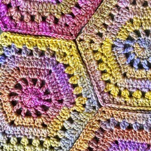 May include: Close-up of a crocheted blanket with a multi-colored pattern of pink, purple, yellow, and orange. The blanket is made of yarn and has a textured surface.