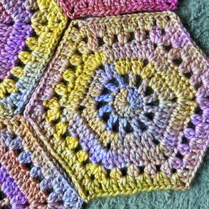 May include: A close-up of a crocheted blanket with a colorful geometric pattern. The blanket is made of yarn in shades of purple, yellow, and pink.