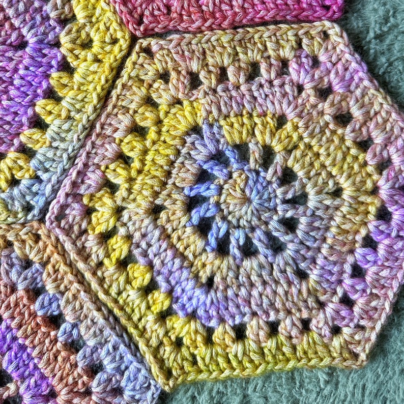 May include: A close-up of a crocheted blanket with a colorful geometric pattern. The blanket is made of yarn in shades of purple, yellow, and pink.