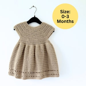 May include: A crocheted baby dress in a light brown colour with a round neckline and short sleeves. The dress has a gathered skirt and a crocheted trim around the bottom. The dress is hanging on a black hanger. The text "Size: 0-3 Months" is visible in the image.