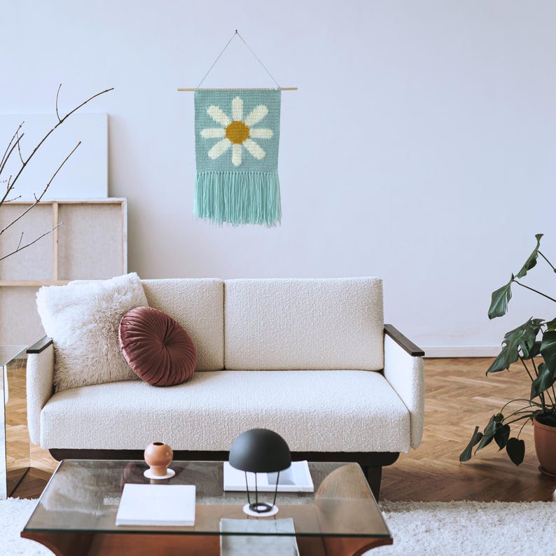 May include: A light blue crocheted wall hanging with a white daisy and a yellow center. The hanging is displayed on a white wall above a white couch with a fluffy white pillow and a burgundy round pillow.