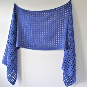 May include: A blue crocheted shawl with a lacy openwork pattern. The shawl is hanging on a white wall.