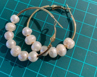 May include: A pair of gold hoop earrings with white pearl accents. The earrings are on a green cutting mat.
