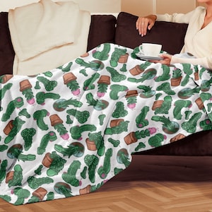 May include: White fleece throw blanket with a repeating pattern of green cactus plants in brown pots. The blanket is folded and draped over a brown couch.