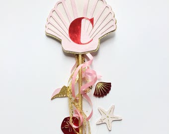 Puede incluir: Una varita de concha de mar de brillo rosa y dorado con la letra "C" en brillo rojo. La varita está decorada con cinta rosa y dorada, una concha rosa, una estrella de mar roja y una estrella de mar dorada.