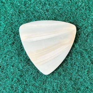 May include: A white, triangular guitar pick with a swirling, layered pattern. The pick is set against a textured, green background, highlighting its shape and design. This accessory is for musicians.
