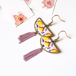 Peut inclure: Une paire de boucles d'oreilles faites à la main avec un motif floral jaune, orange et violet. Les boucles d'oreilles ont une forme de demi-lune avec un cadre en métal doré et un pompon rose pendant. Art floral à l'aquarelle en arrière-plan.