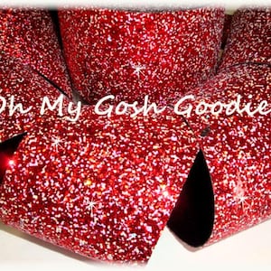 Puede incluir: Cinta roja de purpurina con un acabado brillante e iridiscente. La cinta está doblada en forma de lazo y el texto "Oh My Gosh Goodies" es visible en el centro.