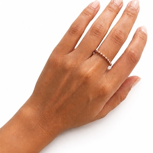 May include: A rose gold beaded ring is worn on a hand with a neutral manicure. The ring features a series of small, evenly spaced beads that encircle the finger. The hand is positioned against a white background, highlighting the ring's design.