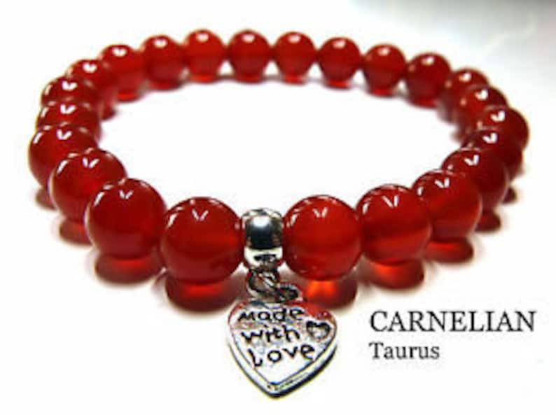 May include: Red carnelian gemstone bracelet with a silver heart charm that says "Made with Love". The bracelet is labeled "CARNELIAN Taurus".