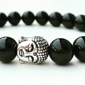 Può includere: Braccialetto con perline nere e ciondolo a testa di Buddha argentato.