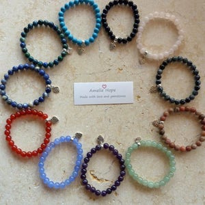 May include: Ten bracelets made with different gemstones, each with a silver heart charm. The bracelets are arranged in a circle on a light brown surface. The text "Amelie Hope Made with love and gemstones" is in the center of the circle.