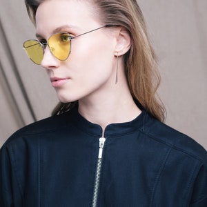 May include: A woman wearing a black collared shirt with a zipper closure and yellow cat-eye sunglasses. She has long brown hair and is wearing thin silver earrings.