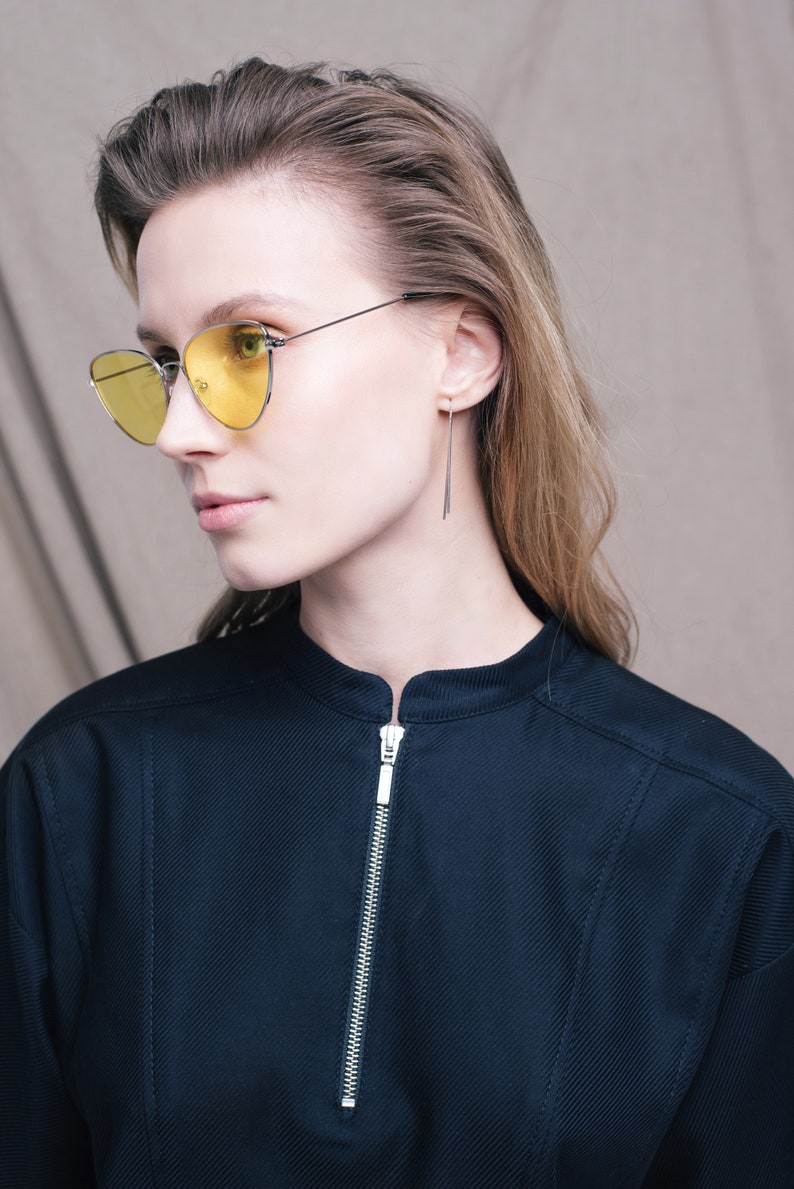 May include: A woman wearing a black collared shirt with a zipper closure and yellow cat-eye sunglasses. She has long brown hair and is wearing thin silver earrings.