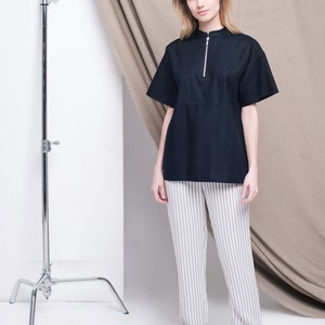 May include: A woman wearing a black short-sleeved shirt with a zipper and white striped pants. She is wearing black sandals and standing in front of a white backdrop with a brown fabric draped behind her.