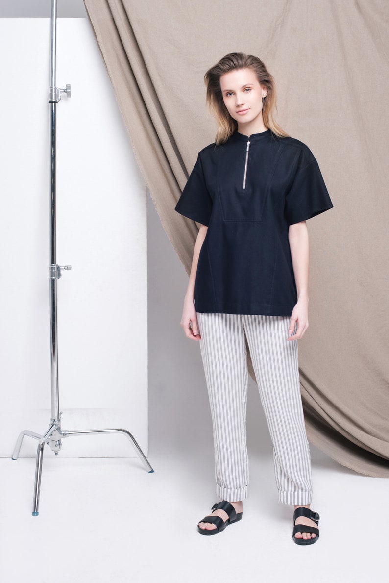 May include: A woman wearing a black short-sleeved shirt with a zipper and white striped pants. She is wearing black sandals and standing in front of a white backdrop with a brown fabric draped behind her.