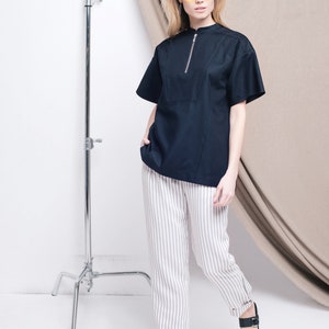 May include: A woman wearing a black short-sleeved shirt with a zipper, white striped pants, and black sandals. She is wearing yellow sunglasses and standing in front of a white backdrop.