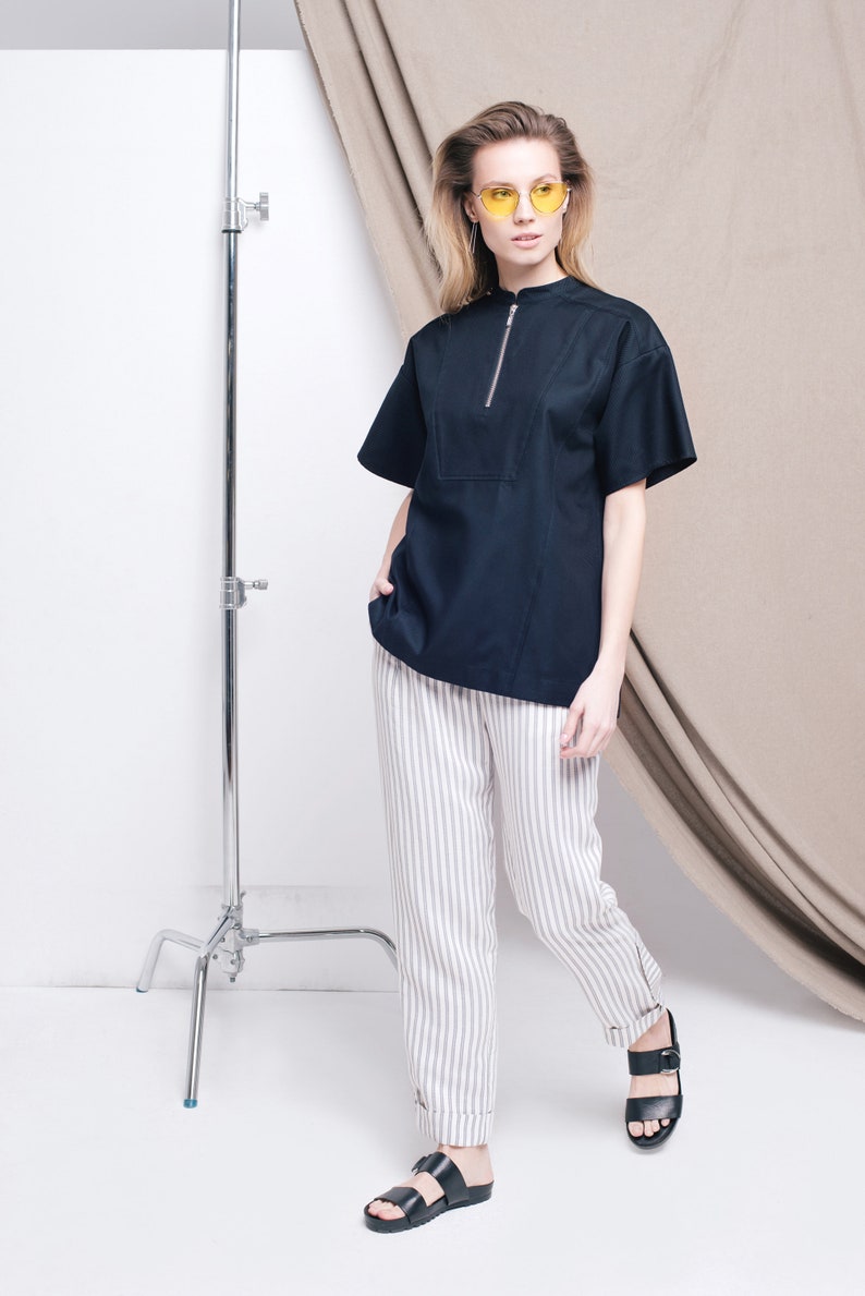 May include: A woman wearing a black short-sleeved shirt with a zipper, white striped pants, and black sandals. She is wearing yellow sunglasses and standing in front of a white backdrop.