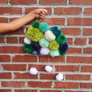 May include: A green, white and blue pom-pom cloud wall hanging with white pom-poms hanging from the bottom.