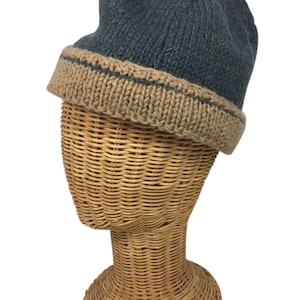 Puede incluir: Un gorro de punto azul verdoso y beige con un borde doblado. El sombrero se exhibe en una cabeza de maniquí de mimbre. El borde es beige con una fina raya oscura. El sombrero parece hecho a mano.