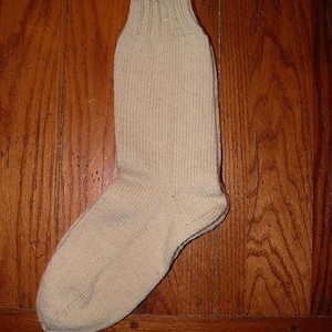Op de afbeelding: Een enkele, beige sok gemaakt van geribbelde gebreide stof. De sok ligt op een houten oppervlak. De sok heeft een versterkte hiel en teen, en een geribbelde boord.