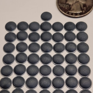 May include: A collection of small, dark gray, circular objects with a textured surface, arranged in a grid pattern. A U.S. quarter coin is placed nearby for size comparison. The coin shows the profile of George Washington and the year 1969.