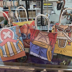 May include: Four colorful tote bags with white handles, each featuring a different cartoon character design. The bags are made of paper and are displayed on a glass surface. The designs include characters with distinctive features.