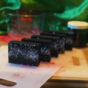 May include: Five rectangular black soap bars with a speckled, crystalline design are arranged on a white cutting board. The soap bars are in a row, with a blurred background of green fabric and a measuring jug.