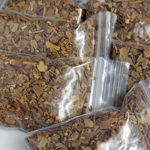 Könnte beinhalten: Mehrere kleine, transparente Plastiktüten gefüllt mit getrocknetem, gehacktem braun-gelbem Pflanzenmaterial. Die Beutel sind leicht überlappend angeordnet. Grüne Blätter und ein hellfarbiger Stein sind im Hintergrund zu sehen.