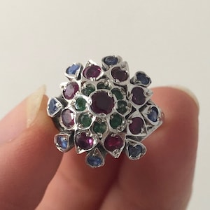 May include: A silver ring featuring a cluster of red, green, and blue gemstones. The ring is set with a variety of small, round stones.