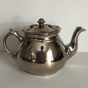 Könnte beinhalten: Eine silberne Teekanne mit einem geriffelten Rand und einer glänzenden Oberfläche. Die Teekanne hat einen kleinen Knopf auf dem Deckel und einen Ausguss an der Seite.