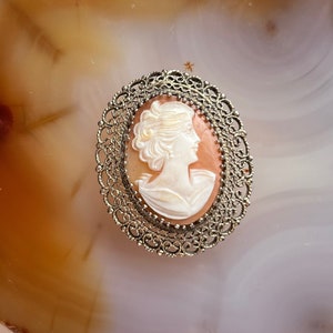 May include: A cameo brooch with a white cameo of a woman's profile set in a silver filigree frame.