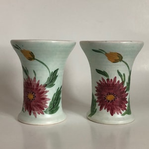 May include: Two small, light blue ceramic vases with a floral design. Each vase features a pink flower with yellow centre and green leaves.