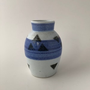 Puede incluir: Un pequeño jarrón de cerámica blanca con un patrón geométrico azul y negro. El jarrón tiene un cuerpo redondeado y un cuello estrecho.