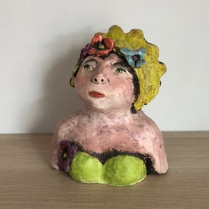 May include: A ceramic bust of a woman with a yellow, flower-shaped hat and a green top. The woman has a pink face and brown eyes. The flowers on her hat are red, purple, and blue.