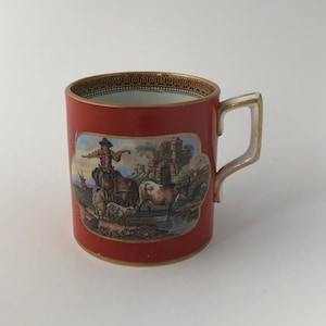 Puede incluir: Una taza de cerámica roja con una escena pastoral que representa a una persona a caballo, ovejas y ganado. La taza tiene un borde y un asa dorados con un acento blanco y dorado. El diseño es de estilo vintage.
