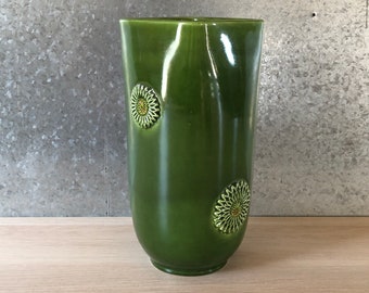 May include: A tall, green ceramic vase with a glossy finish and two raised floral designs. The vase is cylindrical in shape and has a rounded bottom.